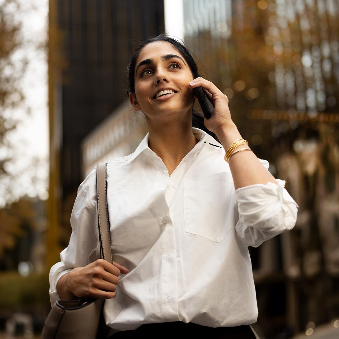A young woman walks through the city. She is talking on her phone and smiling confidently.