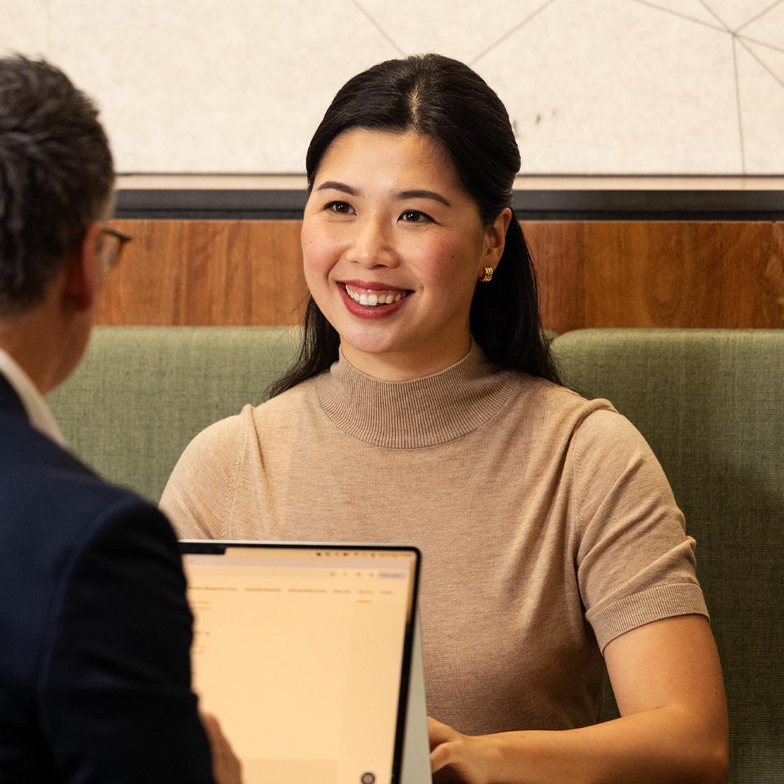 A woman and man sit at a table in a workplace. The man has a laptop, while the woman is smiling.