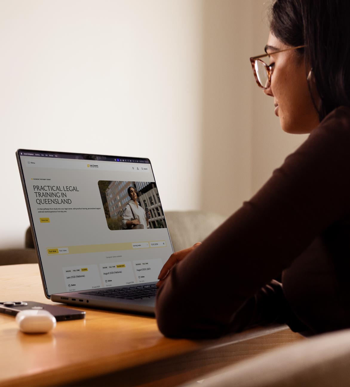 A young woman looks at her laptop sitting at a desk. The screen shows a webpage.